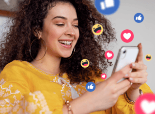 Mulher com cabelos cacheados e blusa amarela sorrindo olhando parao celular om emoticons saindo dele.